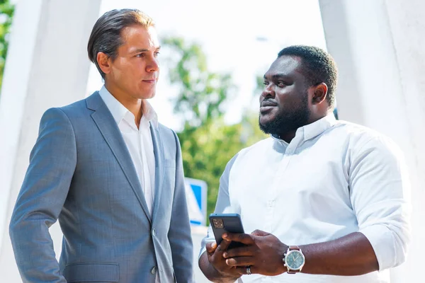 Hombre de negocios afroamericano y su colega frente al moderno edificio de oficinas. Los inversores financieros están hablando al aire libre. Concepto bancario y empresarial. — Foto de Stock