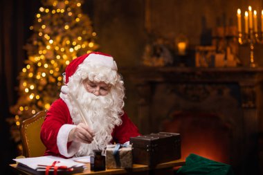 Noel Baba 'nın işyeri. Neşeli Noel Baba masada otururken mektubu yazıyor. Arka planda şömine ve Noel ağacı var. Noel konsepti.