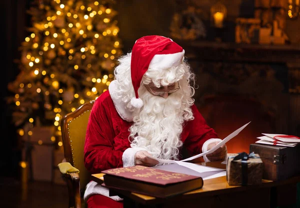 Lieu de travail du Père Noël. Le Père Noël joyeux lit les lettres des enfants assis à la table. Cheminée et sapin de Noël en arrière-plan. Concept de Noël. — Photo