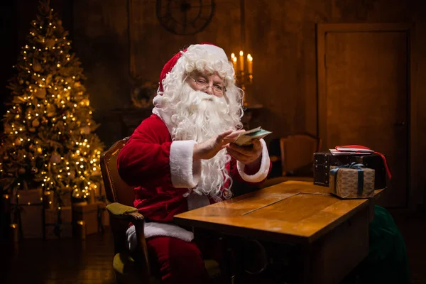 サンタクロースの職場。陽気なサンタはテーブルに座ってお金を数えている。背景にある暖炉とクリスマスツリー。クリスマスのコンセプト. — ストック写真