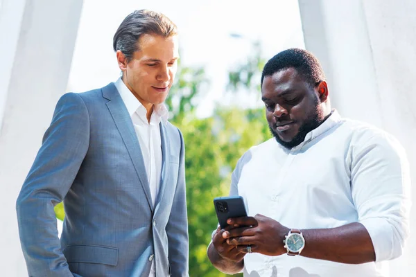 Uomo d'affari afro-americano e il suo collega di fronte a un moderno edificio per uffici. Gli investitori finanziari stanno parlando all'aperto. Concetto bancario e aziendale. Immagini Stock Royalty Free