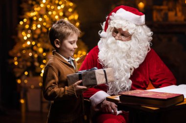 Noel Baba ve küçük çocuk. Neşeli Noel Baba masada otururken çalışıyor. Arka planda şömine ve Noel ağacı var. Noel konsepti.