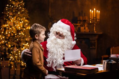 Noel Baba ve küçük çocuk. Neşeli Noel Baba masada otururken çalışıyor. Arka planda şömine ve Noel ağacı var. Noel konsepti.