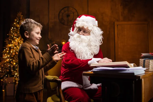 A Mikulás és a kisfiú. Vidám Mikulás dolgozik, miközben ül az asztalnál. Kandalló és karácsonyfa a háttérben. Karácsonyi koncepció. — Stock Fotó