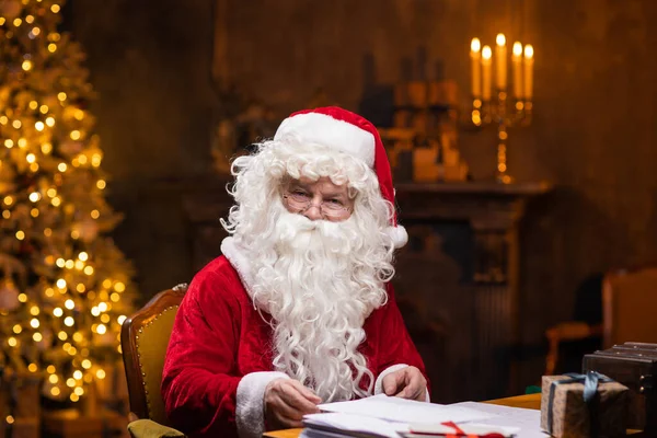 Χώρος εργασίας του Άγιου Βασίλη. Ο χαρούμενος Άγιος Βασίλης δουλεύει ενώ κάθεται στο τραπέζι. Τζάκι και χριστουγεννιάτικο δέντρο στο παρασκήνιο. Χριστουγεννιάτικη ιδέα. — Φωτογραφία Αρχείου