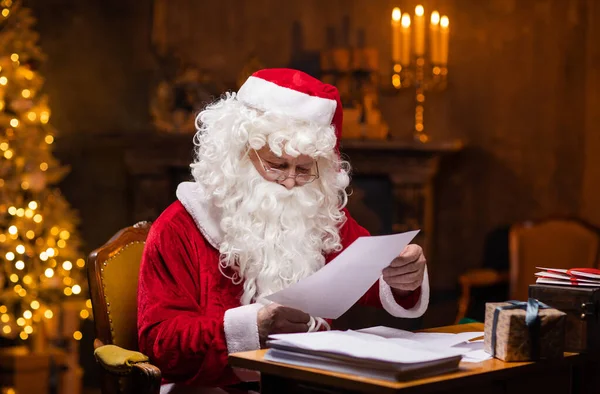 サンタクロースの職場。陽気サンタはテーブルに座っている間に子供たちからの手紙を読んでいます。背景にある暖炉とクリスマスツリー。クリスマスのコンセプト. — ストック写真