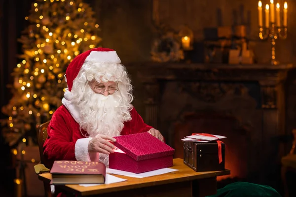 Arbeitsplatz des Weihnachtsmannes. Gut gelaunt zaubert der Weihnachtsmann am Tisch über die Geschenkbox. Kamin und Weihnachtsbaum im Hintergrund. Weihnachtskonzept. — Stockfoto