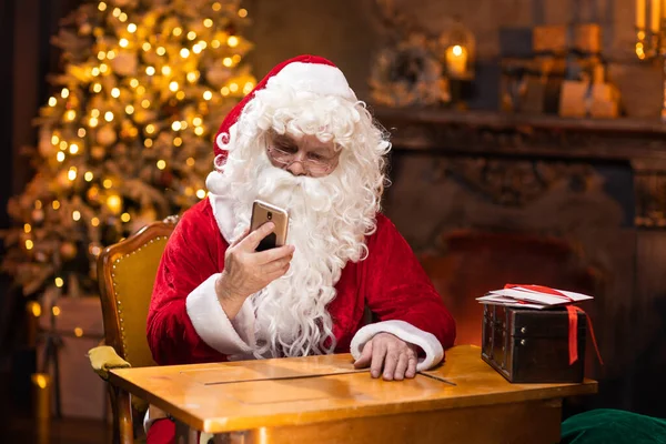 Workplace of Santa Claus. Cheerful Santa is working using smartphone while sitting at the table. Fireplace and Christmas Tree in the background. Christmas concept. — Stock Photo, Image