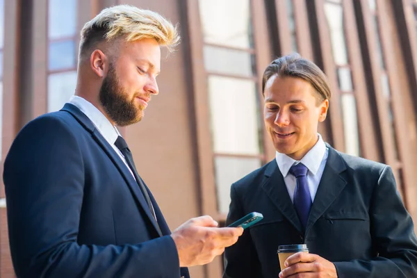 Confident businessman and his colleague are using smartphone in front of modern office building. Financial investors are talking outdoor. Banking and business. Royalty Free Stock Photos