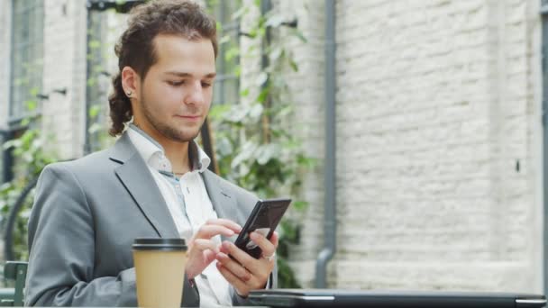 Unge succesfulde kaukasiske forretningsmand arbejder i udendørs cafe ved hjælp af en smartphone. Forretning, freelance og fjernarbejde koncept. – Stock-video