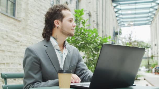 Joven empresario caucásico exitoso trabaja en una cafetería al aire libre con un ordenador y un teléfono inteligente. Negocios, freelance y trabajo remoto. — Vídeos de Stock