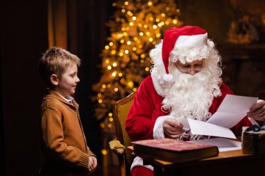 Noel Baba ve küçük çocuk. Neşeli Noel Baba masada otururken çalışıyor. Arka planda şömine ve Noel ağacı var. Noel konsepti.