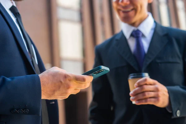 Der selbstbewusste Geschäftsmann und sein Kollege bedienen sich vor modernen Bürogebäuden des Smartphones. Finanzinvestoren reden im Freien. Banken und Unternehmen. — Stockfoto