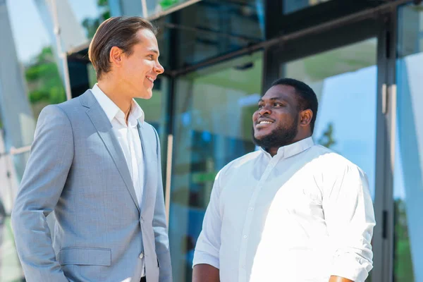 Uomo d'affari afro-americano e il suo collega di fronte a un moderno edificio per uffici. Gli investitori finanziari stanno parlando all'aperto. Concetto bancario e aziendale. — Foto Stock