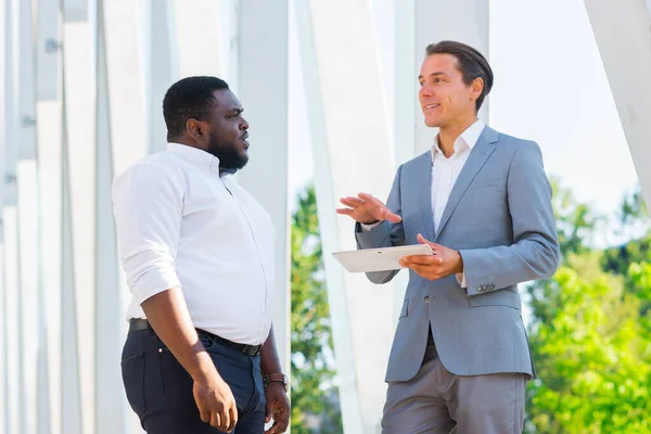 Hombre de negocios afroamericano y su colega frente al moderno edificio de oficinas. Los inversores financieros están hablando al aire libre. Concepto bancario y empresarial. — Foto de Stock