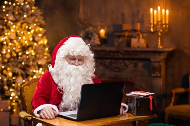 Noel Baba 'nın işyeri. Neşeli Noel Baba masada otururken dizüstü bilgisayarda çalışıyor. Arka planda şömine ve Noel ağacı var. Noel konsepti.