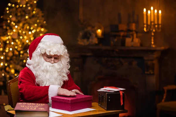 サンタクロースの職場 陽気サンタはテーブルに座っている間にギフトボックスの上に集まっています 背景にある暖炉とクリスマスツリー 伝統的なクリスマスのコンセプト — ストック写真