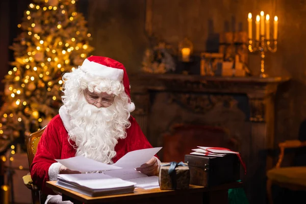 Noel Baba 'nın işyeri. Neşeli Noel Baba masada otururken çocuklardan gelen mektupları okuyor. Arka planda şömine ve Noel ağacı var. Noel konsepti. — Stok fotoğraf