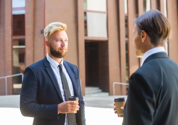 Homme d'affaires confiant et son collègue devant un immeuble de bureaux moderne. Les investisseurs financiers parlent en plein air. Banques et entreprises. — Photo