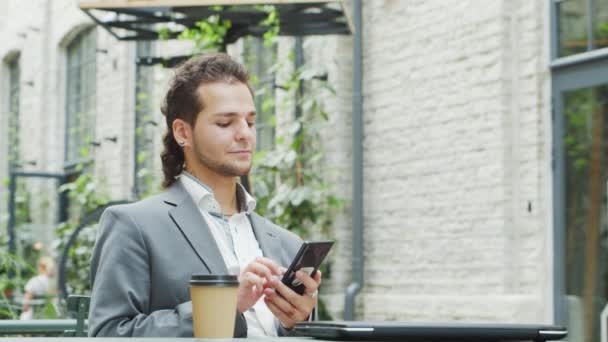 Młody, odnoszący sukcesy biały biznesmen pracuje w kawiarni na świeżym powietrzu za pomocą smartfona. Koncepcja biznesu, pracy na własny rachunek i pracy na odległość. — Wideo stockowe
