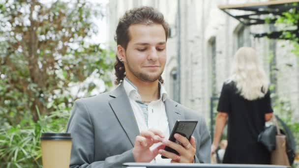 Jonge succesvolle Kaukasische zakenman werkt in outdoor cafe met behulp van een smartphone. Bedrijfs-, freelance- en telewerkconcept. — Stockvideo