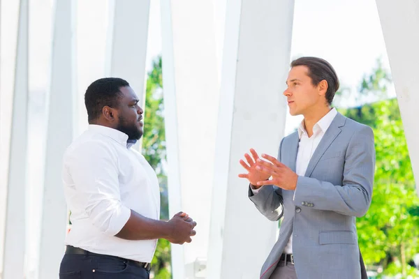 Empresário afro-americano e seu colega em frente ao edifício de escritórios moderno. Os investidores financeiros estão a falar ao ar livre. Conceito bancário e empresarial. — Fotografia de Stock