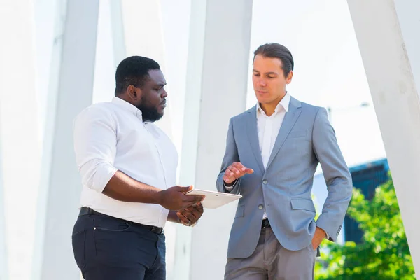 Empresário afro-americano e seu colega em frente ao edifício de escritórios moderno. Os investidores financeiros estão a falar ao ar livre. Conceito bancário e empresarial. — Fotografia de Stock