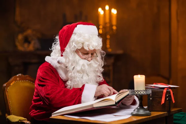 サンタクロースの職場。陽気サンタはテーブルに座っている間に願い事の本を読んでいます。背景にある暖炉とクリスマスツリー。クリスマスのコンセプト. — ストック写真