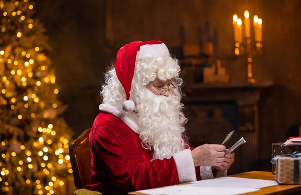 Χώρος εργασίας του Άγιου Βασίλη. Ο χαρούμενος Άγιος Βασίλης μετράει λεφτά ενώ κάθεται στο τραπέζι. Τζάκι και χριστουγεννιάτικο δέντρο στο παρασκήνιο. Χριστουγεννιάτικη ιδέα. — Φωτογραφία Αρχείου