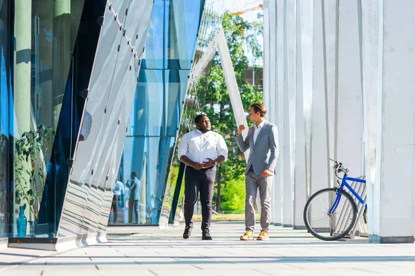 Hombre de negocios afroamericano y su colega frente al moderno edificio de oficinas. Los inversores financieros están hablando al aire libre. Concepto bancario y empresarial. — Foto de Stock