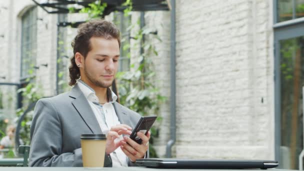 Joven empresario caucásico exitoso trabaja en la cafetería al aire libre con un teléfono inteligente. Concepto de trabajo empresarial, autónomo y remoto. — Vídeos de Stock