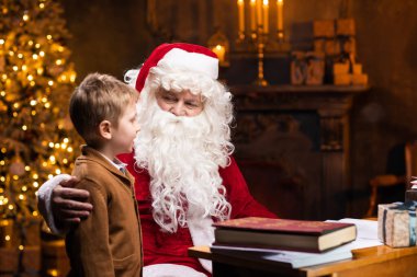 Noel Baba ve küçük çocuk. Neşeli Noel Baba masada otururken çalışıyor. Arka planda şömine ve Noel ağacı var. Noel konsepti.