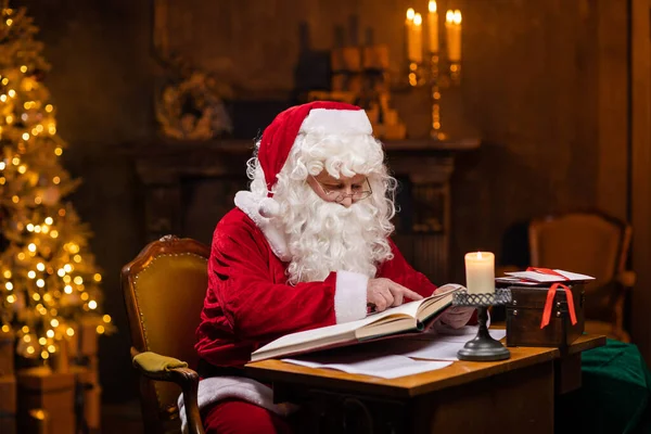 Workplace of Santa Claus. Cheerful Santa is reading the book of wishes while sitting at the table. Fireplace and Christmas Tree in the background. Christmas concept.