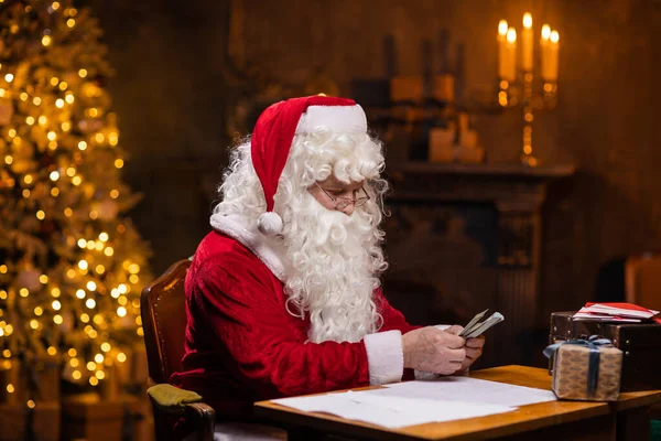 Workplace of Santa Claus. Cheerful Santa is counting money while sitting at the table. Fireplace and Christmas Tree in the background. Christmas concept. — Stock Photo, Image