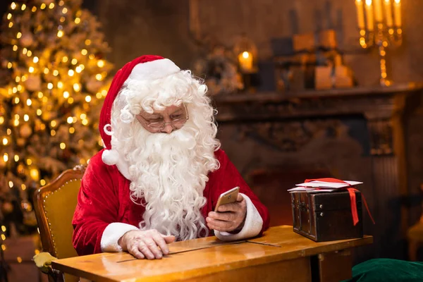 Arbeitsplatz des Weihnachtsmannes. Der fröhliche Weihnachtsmann arbeitet mit dem Smartphone, während er am Tisch sitzt. Kamin und Weihnachtsbaum im Hintergrund. Weihnachtskonzept. — Stockfoto