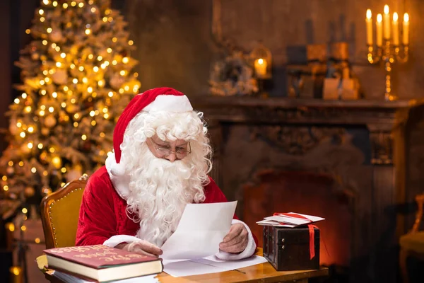 サンタクロースの職場。陽気サンタはテーブルに座っている間に子供たちからの手紙を読んでいます。背景にある暖炉とクリスマスツリー。クリスマスのコンセプト. — ストック写真