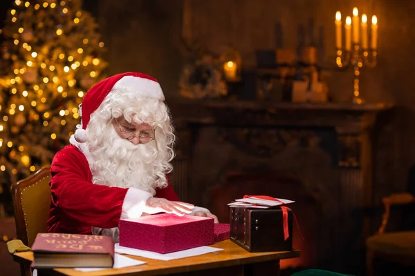 Arbeitsplatz des Weihnachtsmannes. Gut gelaunt zaubert der Weihnachtsmann am Tisch über die Geschenkbox. Kamin und Weihnachtsbaum im Hintergrund. Weihnachtskonzept. — Stockfoto