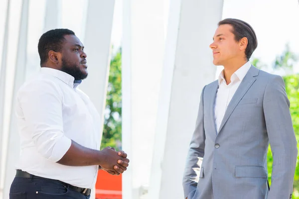 Uomo d'affari afro-americano e il suo collega di fronte a un moderno edificio per uffici. Gli investitori finanziari stanno parlando all'aperto. Concetto bancario e aziendale. — Foto Stock