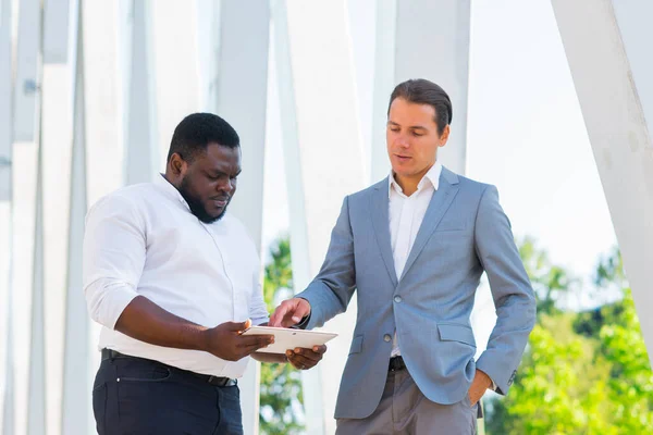 Uomo d'affari afro-americano e il suo collega di fronte a un moderno edificio per uffici. Gli investitori finanziari stanno parlando all'aperto. Concetto bancario e aziendale. — Foto Stock