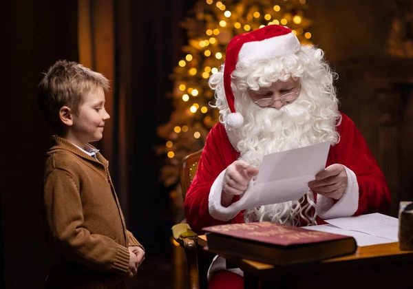 Père Noël et petit garçon. Père Noël joyeux travaille tout en étant assis à la table. Cheminée et sapin de Noël en arrière-plan. Concept de Noël. — Photo