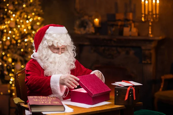 Noel Baba 'nın işyeri. Neşeli Noel Baba masada otururken hediye kutusunun üzerinde hokkabazlık yapıyor. Arka planda şömine ve Noel ağacı var. Noel konsepti. — Stok fotoğraf