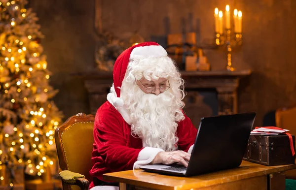 Lugar de trabajo de Santa Claus. Alegre Santa está trabajando en el portátil mientras está sentado en la mesa. Chimenea y árbol de Navidad en el fondo. Concepto de Navidad. —  Fotos de Stock