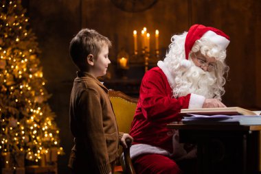Noel Baba ve küçük çocuk. Neşeli Noel Baba masada otururken çalışıyor. Arka planda şömine ve Noel ağacı var. Noel konsepti.