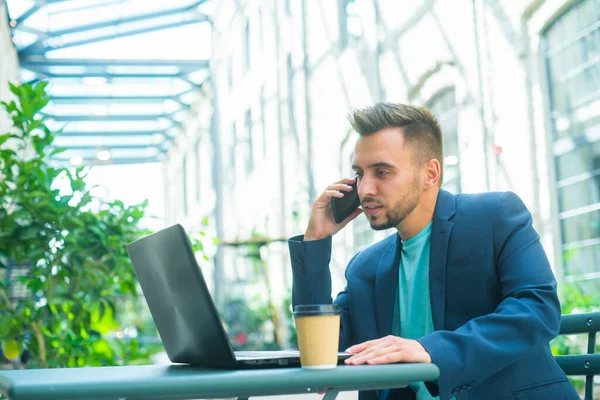 Jonge succesvolle Kaukasische zakenman werkt in een outdoor cafe met behulp van een computer. Bedrijven, freelance en werk op afstand. — Stockfoto