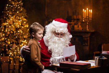 Noel Baba ve küçük çocuk. Neşeli Noel Baba masada otururken çalışıyor. Arka planda şömine ve Noel ağacı var. Noel konsepti.