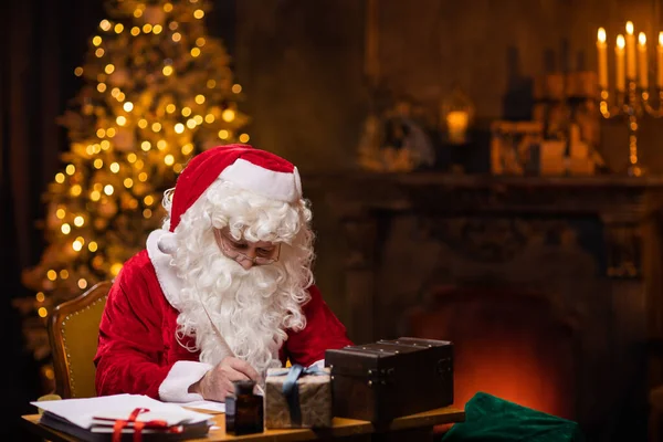 Workplace of Santa Claus. Cheerful Santa is writing the letter while sitting at the table. Fireplace and Christmas Tree in the background. Christmas concept.