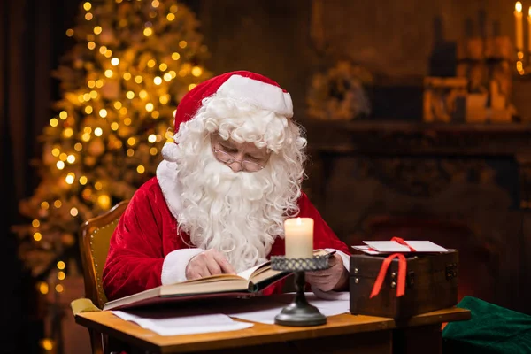 Workplace of Santa Claus. Cheerful Santa is reading the book of wishes while sitting at the table. Fireplace and Christmas Tree in the background. Christmas concept. — Stock Photo, Image