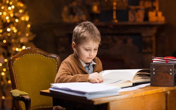 Мальчик читает волшебную книгу, сидя за столом. Интерьер дома с елкой и камином. Традиционная рождественская идея. — стоковое фото