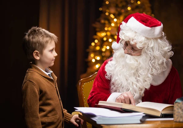 Noel Baba ve küçük çocuk. Neşeli Noel Baba masada otururken çalışıyor. Arka planda şömine ve Noel ağacı var. Noel konsepti. — Stok fotoğraf