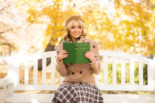 Schöne lächelnde junge Frau, die im Herbst auf der Bank sitzt und ein neues Buch liest — Stockfoto